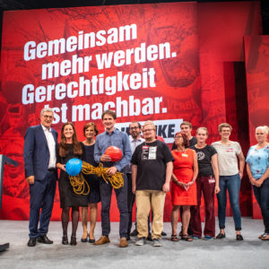 LINKE Mitglieder stehen vor einer Parteitagswand auf der in großen Lettern "Gemeinsam mehr werden" steht.